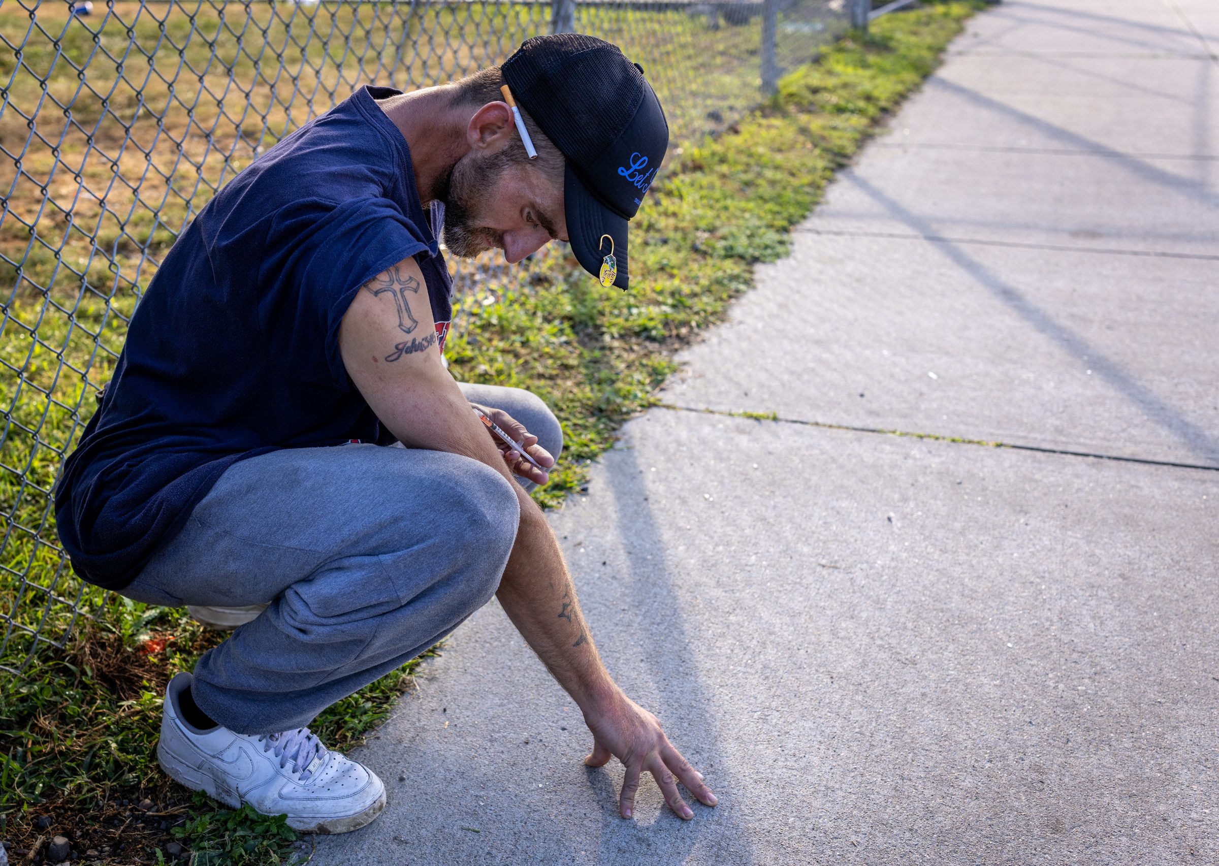 A man crouches in the narrow strip of grass between a chain-link fence and an empty sidewalk at Mass and Cass, a syringe in one hand and the other hand flat on the concrete