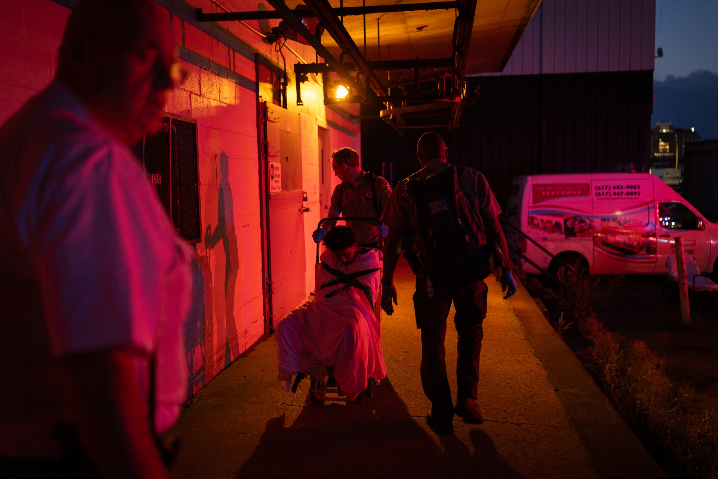Under red emergency lighting, paramedics wheel a woman wrapped in a blanket along a loading dock wall toward an ambulance