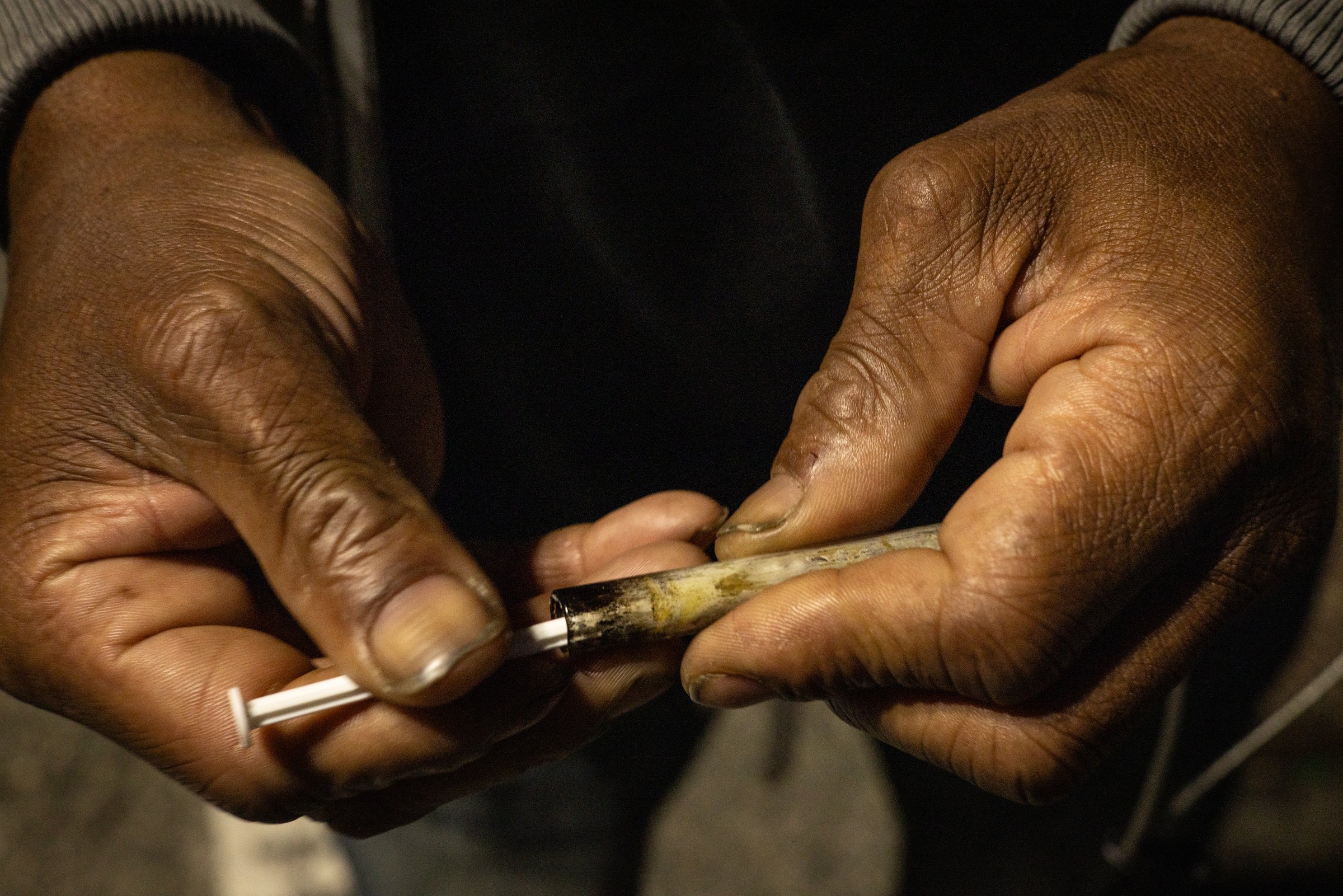 Two worn hands hold a used glass pipe and a thin packing stick, Mass and Cass
